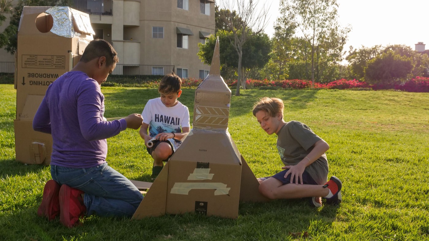 Three campers kneeling on grass building a tall cardboard rocket together