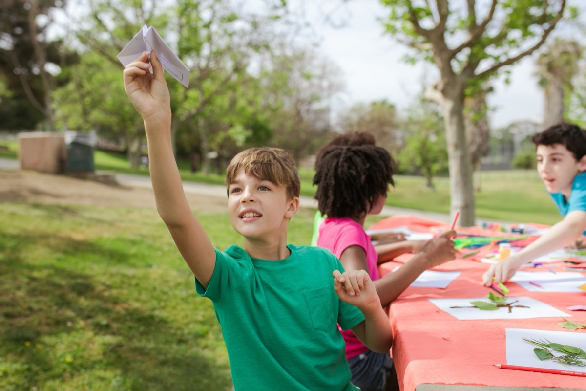 Campers at an outdoor table making things, one holding up a paper aeroplane