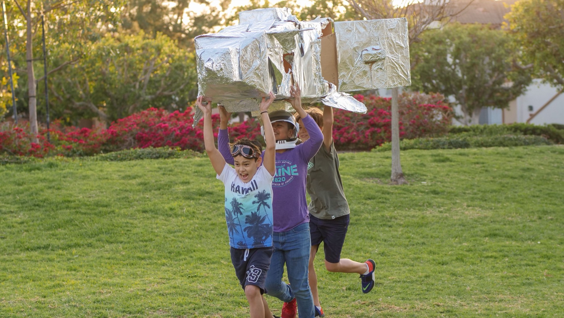 Three campers running across a field, grinning, carrying a large foil covered rocket they built above their heads