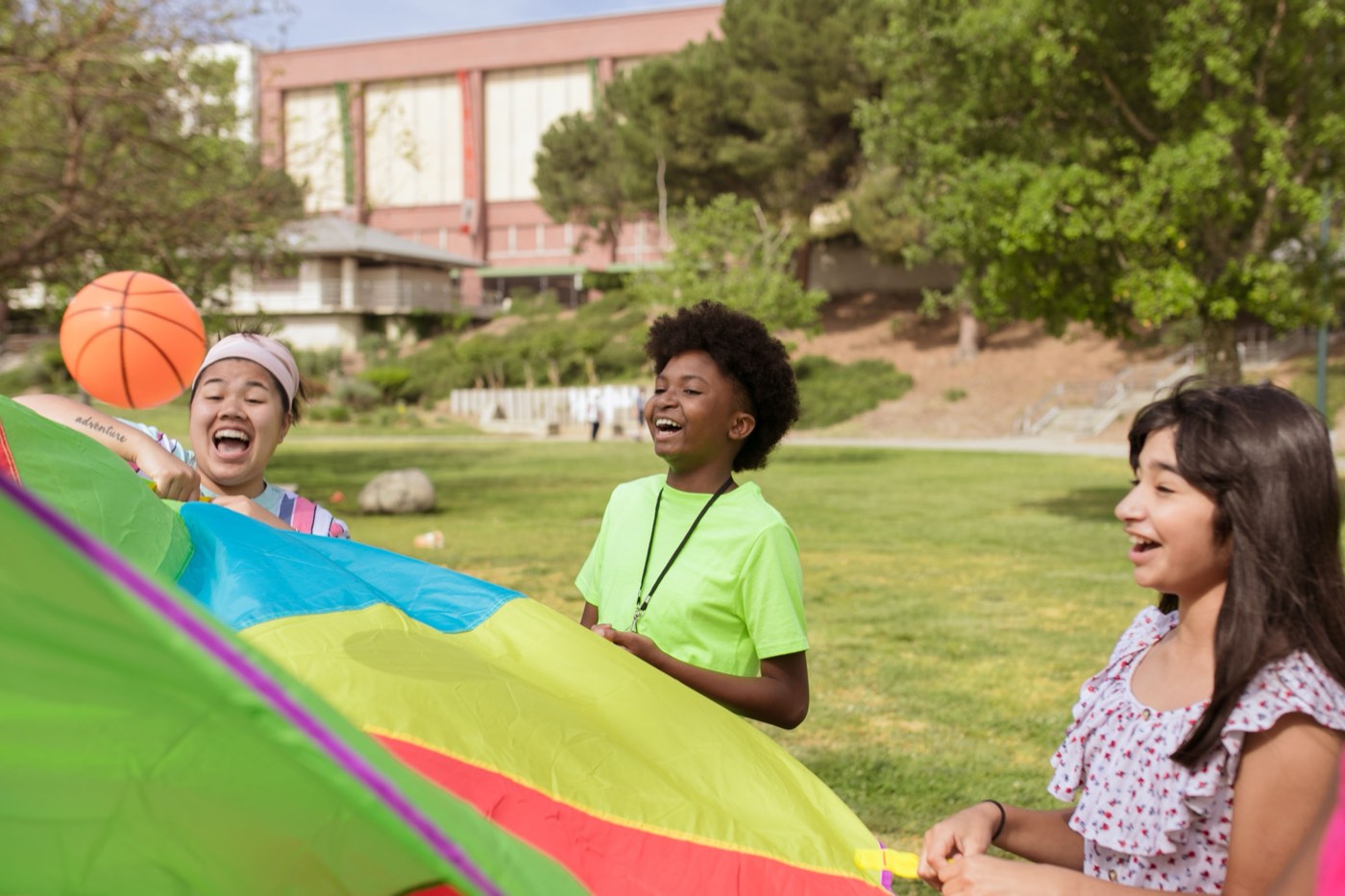 Campers laughing while bouncing a basketball on a colourful parachute in a park